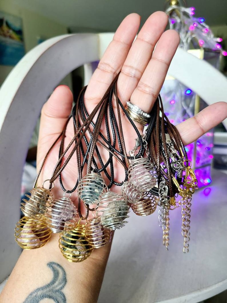 May include: A collection of necklaces with caged crystal pendants. The pendants are made of silver and gold wire and contain various types of crystals. The necklaces are made of brown cord.