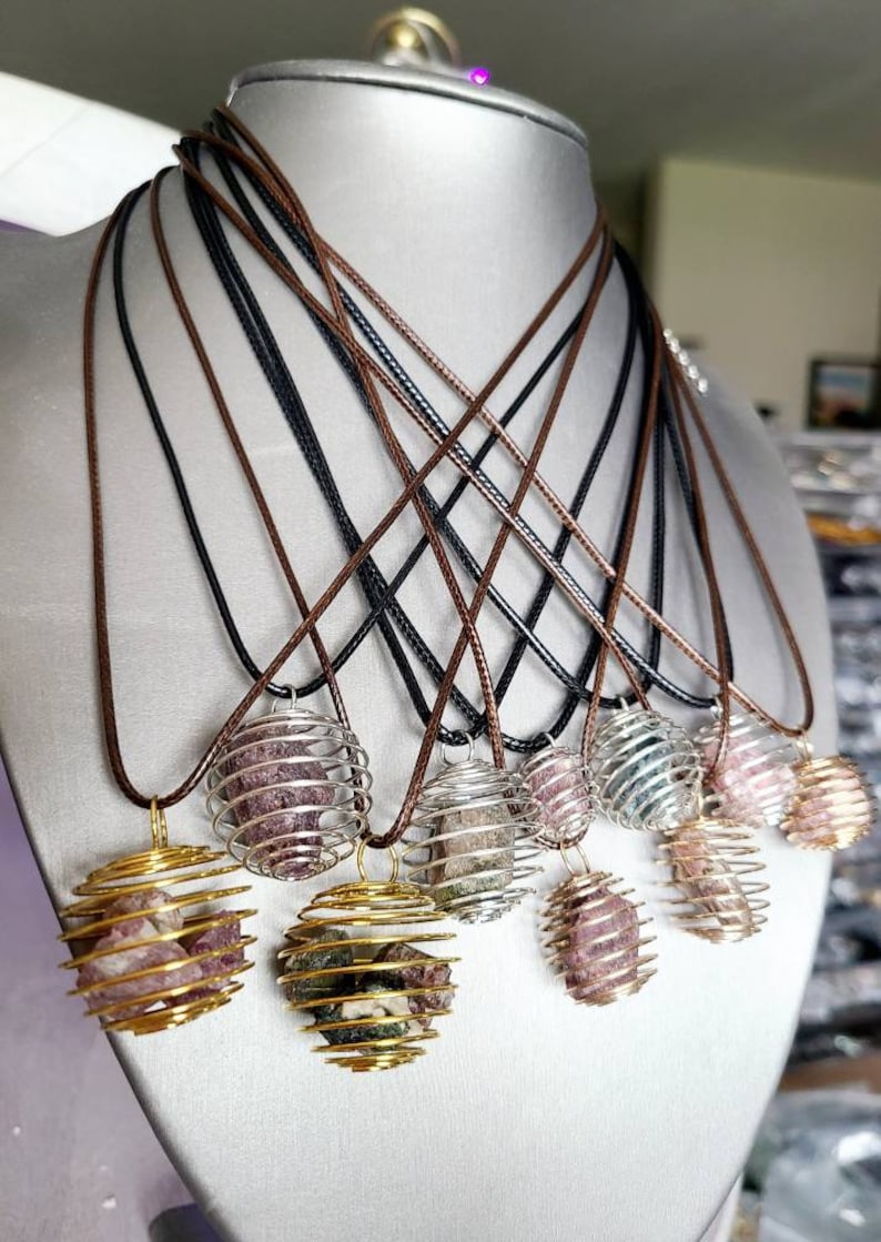 May include: A set of eight necklaces with crystal cage pendants. Each pendant features a different colored crystal encased in a gold or silver wire cage. The necklaces are displayed on a mannequin and have brown and black cords.