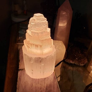 Op de afbeelding: Een witte selenietkristal lamp in de vorm van een toren, verlicht en gloeiend, op een houten oppervlak met andere kristallen.