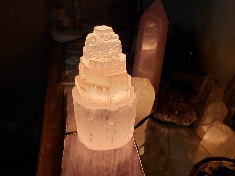 Op de afbeelding: Een witte selenietkristal lamp in de vorm van een toren, verlicht en gloeiend, op een houten oppervlak met andere kristallen.