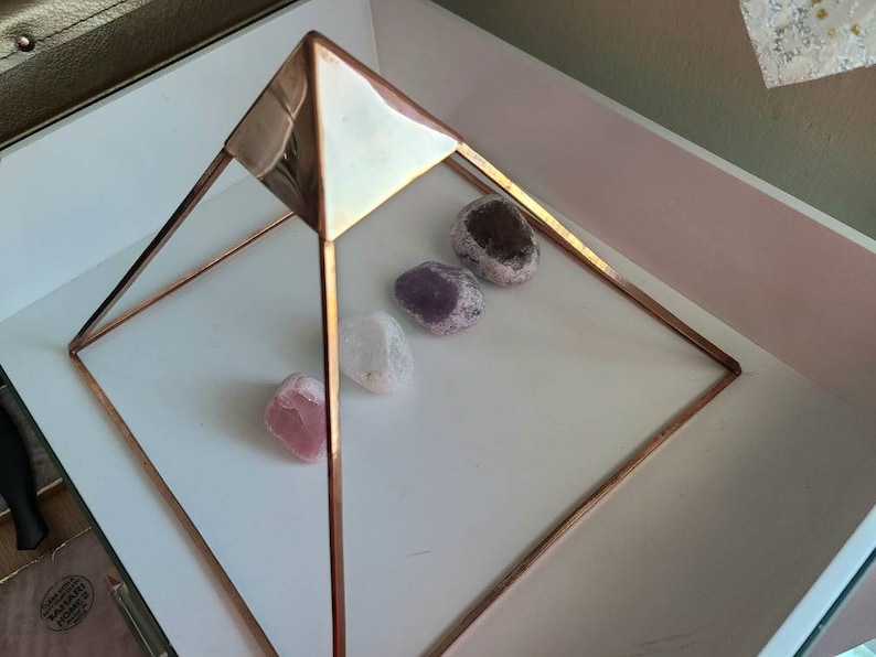 May include: A copper wire pyramid with four smooth, rounded stones inside. The stones are various shades of purple and pink.