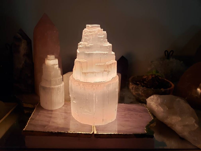 Op de afbeelding: Een witte selenietkristal lamp op een roze en gouden basis. De lamp is piramidevormig en wordt van binnenuit verlicht, waardoor een warme gloed ontstaat.