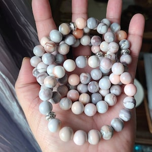 Op de afbeelding: Een set van twee armbanden gemaakt met roze, witte en grijze agaat kralen. De kralen zijn rond en hebben een natuurlijke, gepolijste afwerking. De armbanden zijn versierd met zilveren metalen kralen.
