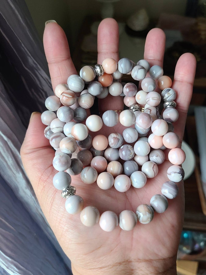 Op de afbeelding: Een set van twee armbanden gemaakt met roze, witte en grijze agaat kralen. De kralen zijn rond en hebben een natuurlijke, gepolijste afwerking. De armbanden zijn versierd met zilveren metalen kralen.