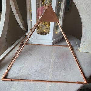May include: A copper pyramid with a triangular top. The pyramid is made of thin copper wire and is sitting on a light wood surface.