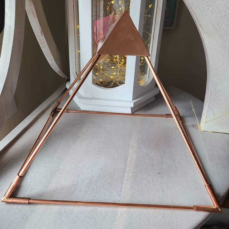 May include: A copper pyramid with a triangular top. The pyramid is made of thin copper wire and is sitting on a light wood surface.