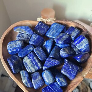 May include: A heart-shaped wooden bowl filled with tumbled lapis lazuli stones. The stones are a deep blue with white veins and have a smooth, polished finish.