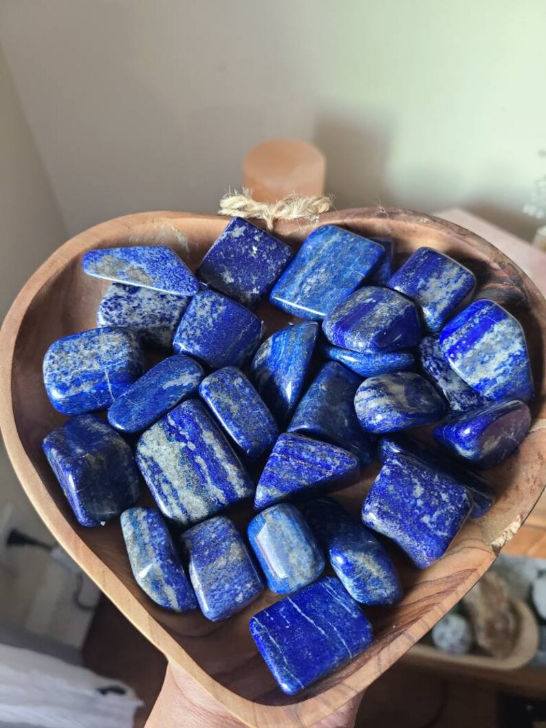 May include: A heart-shaped wooden bowl filled with tumbled lapis lazuli stones. The stones are a deep blue with white veins and have a smooth, polished finish.