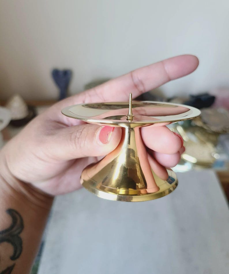 Pu&ograve; includere: Un piccolo portacandele dorato con base rotonda e stelo svasato e conico. La parte superiore presenta una superficie circolare piatta con una punta centrale per tenere una candela. Il portacandele &egrave; in metallo lucido.