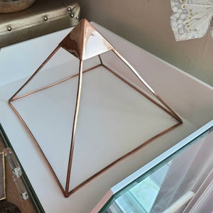 May include: A rose gold metal pyramid shaped crystal grid or stand. The pyramid is on a white surface.