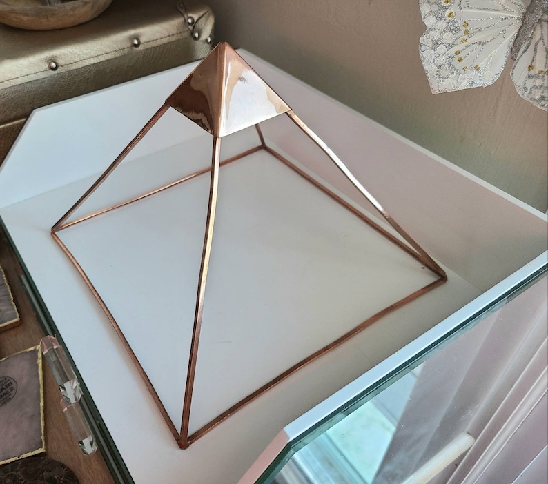 May include: A rose gold metal pyramid shaped crystal grid or stand. The pyramid is on a white surface.