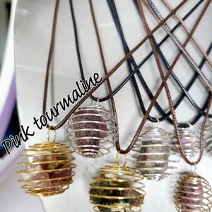 May include: Five necklaces with different colored gemstones encased in gold and silver wire cages. The necklaces are on a brown surface. The gemstones are pink tourmaline and green tourmaline.