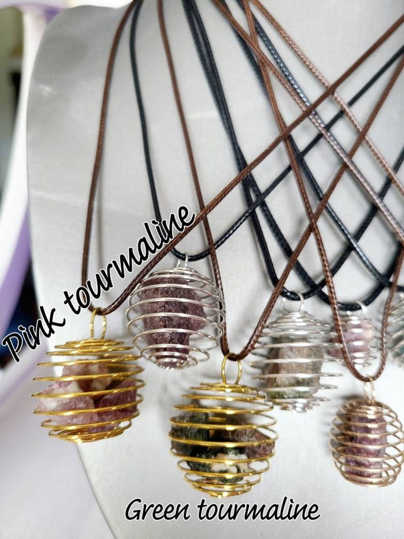 May include: Five necklaces with different colored gemstones encased in gold and silver wire cages. The necklaces are on a brown surface. The gemstones are pink tourmaline and green tourmaline.