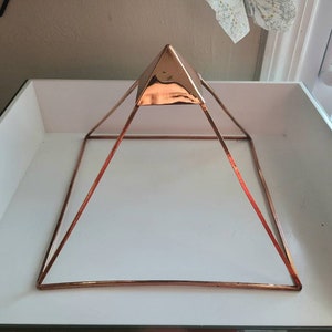 May include: A rose gold metal pyramid with a square base. The pyramid is sitting on a white surface.