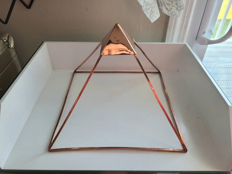 May include: A rose gold metal pyramid with a square base. The pyramid is sitting on a white surface.