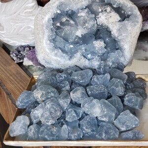 Peut inclure: Une grande g&eacute;ode de c&eacute;lestine bleu clair avec un ext&eacute;rieur gris rugueux et un int&eacute;rieur cristallin, plac&eacute;e derri&egrave;re une collection de nombreuses petites pierres bleu clair translucides et polies sur un plateau rectangulaire.