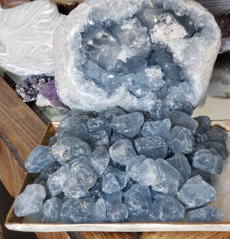 Peut inclure: Une grande g&eacute;ode de c&eacute;lestine bleu clair avec un ext&eacute;rieur gris rugueux et un int&eacute;rieur cristallin, plac&eacute;e derri&egrave;re une collection de nombreuses petites pierres bleu clair translucides et polies sur un plateau rectangulaire.