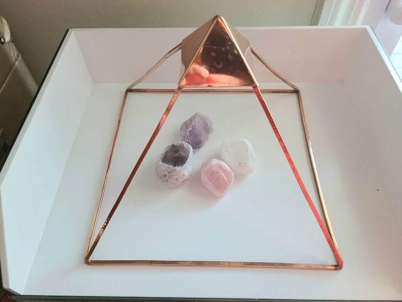 May include: A copper-toned metal pyramid with four sides and a square base. The pyramid is sitting on a white surface and contains three crystals: a purple amethyst, a pink rose quartz, and a clear quartz.