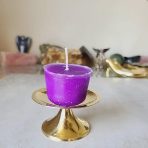 Pu&ograve; includere: Una vivace candela viola con uno stoppino bianco &egrave; posta su un piccolo piedistallo in metallo dorato. La candela ha una superficie liscia ed &egrave; circondata da altri oggetti decorativi sullo sfondo.