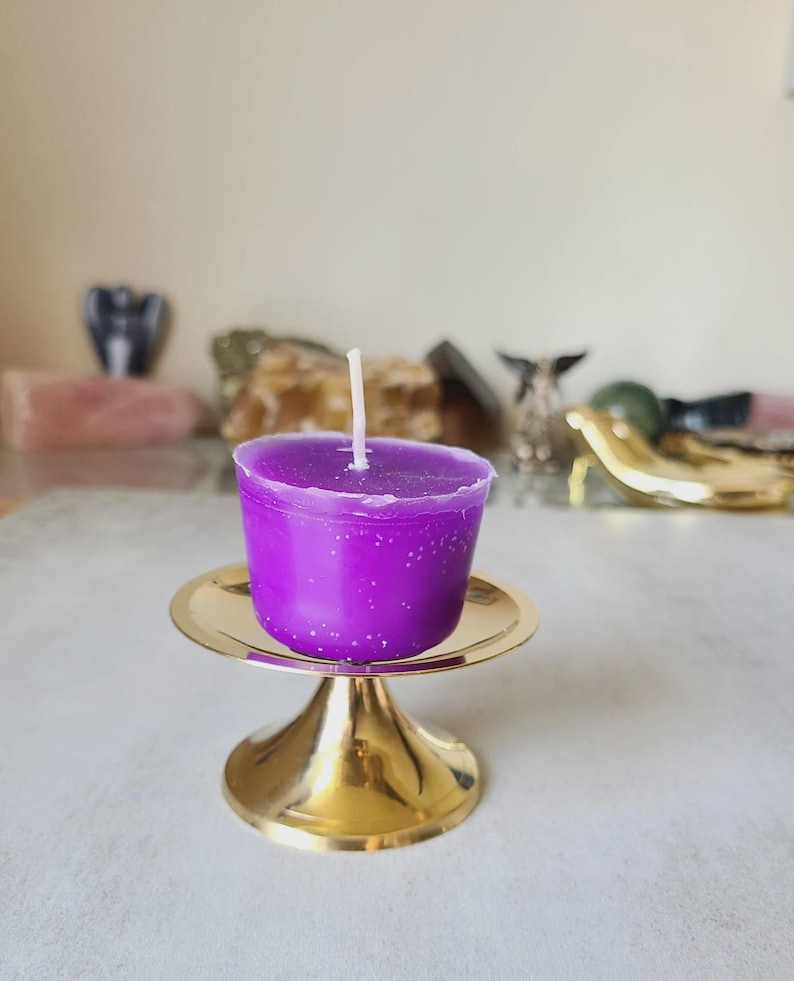 Pu&ograve; includere: Una vivace candela viola con uno stoppino bianco &egrave; posta su un piccolo piedistallo in metallo dorato. La candela ha una superficie liscia ed &egrave; circondata da altri oggetti decorativi sullo sfondo.