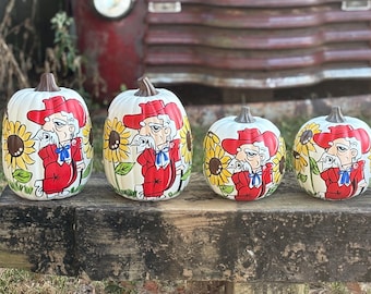 May include: Four white decorative pumpkins featuring a hand-painted illustration of a man with a white mustache wearing a red hat and red suit, accompanied by yellow sunflowers. Each pumpkin is topped with a brown stem.