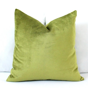 May include: A square, olive green velvet pillow. The pillow is a solid color and has a soft, plush appearance. The pillow is placed on a white surface.