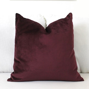 May include: A square throw pillow featuring a deep burgundy velvet fabric cover. The pillow has a soft, plush texture with a slight sheen across the surface, showcasing a solid, dark red color throughout the entire front face of the cushion.
