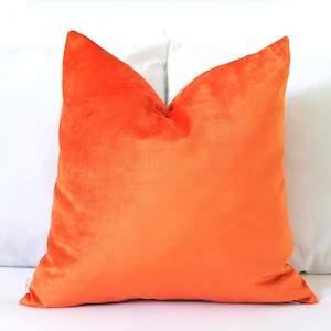 May include: A square cushion in a vivid orange velvet. The cushion has a soft, plush texture and is a bright shade of orange. It is placed on a white surface, with a white cushion visible behind it.
