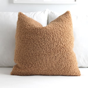May include: A square, tan-colored pillow with a textured, plush surface. The pillow is made of a soft, fuzzy material and is placed on a white surface. The pillow is a decorative item for a bed or couch.