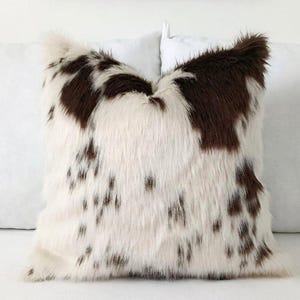 May include: A square decorative pillow with a faux fur cover. The pillow features a white base with brown spots and patches, creating a cowhide pattern. The pillow is fluffy and soft, perfect for adding texture to a living space.