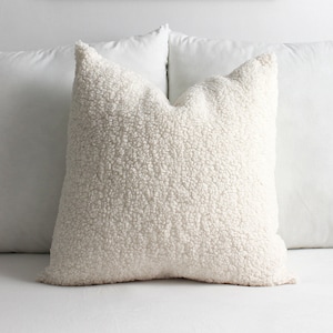 May include: A fluffy, off-white decorative pillow with a textured, sherpa-like surface. The pillow is square-shaped and rests on a white surface, suggesting a bed or couch setting. The soft, inviting texture adds a cozy element to the room.