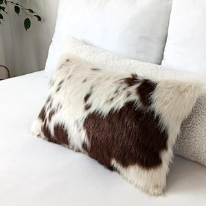 May include: A rectangular throw pillow featuring a faux fur cover with a brown and white cow print pattern. The pillow has a soft, textured appearance and is positioned in front of a white sherpa-style pillow and plain white bedding.