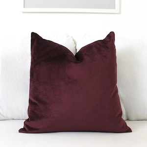 May include: A square throw cushion cover in a deep, solid burgundy colour. The fabric has a soft, velvet-like texture with a slight sheen that catches the light, creating subtle variations in the dark red tone across the surface of the cushion.