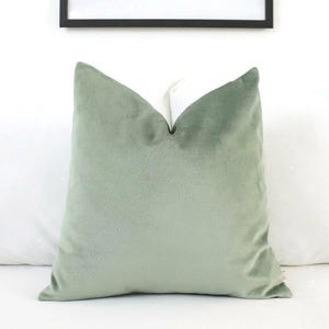 May include: A square throw pillow cover in a solid sage green color. The fabric has a soft, velvet-like texture with a slight sheen. The pillow is plumped to create a subtle indentation at the top center, showcasing the plush, smooth material.