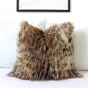 Op de afbeelding: Een vierkant sierkussen met een hoes van imitatiebont met lange haren in een gevarieerd patroon van bruine, beige en crèmekleurige tinten. De ruige textuur zorgt voor een dichte, gelaagde uitstraling over het gehele oppervlak van het kussen.