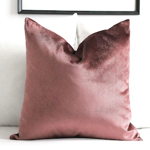 May include: A square throw pillow featuring a solid mauve-toned velvet fabric with a soft, light-reflecting sheen. The pillow has a plush, filled appearance with crisp, defined edges and a smooth, uniform texture across the entire front surface.