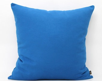May include: A square throw cushion in a solid, vibrant blue colour. The cushion features a smooth, uniform fabric texture across its entire surface. A small, black rectangular tag with the letters SN is attached to the bottom right corner edge.