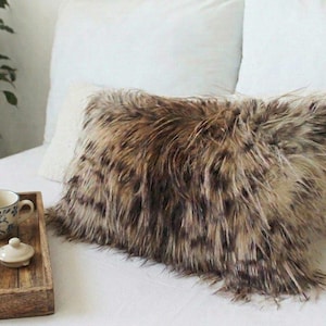 Könnte beinhalten: Ein rechteckiges Kissen aus Kunstfell in Braun- und Beigetönen liegt auf einem weißen Bett. Das Kissen hat eine zottelige Textur und liegt vor weißen Kissen. Ein Holztablett mit einer Tasse und Untertasse befindet sich im Vordergrund.