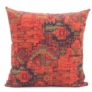 May include: A square throw pillow featuring a woven fabric with a complex geometric pattern in shades of vibrant red, deep charcoal black, and muted olive green. The design consists of intricate interlocking lines, angular shapes, and motifs.