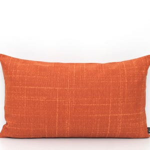 May include: A rectangular throw pillow in a vibrant burnt orange hue, featuring a subtle, light-colored grid pattern of thin, intersecting lines across the fabric surface. A small, rectangular white brand tag is visible on the bottom right corner.