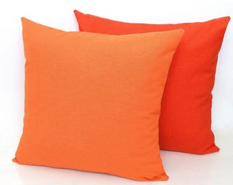 Op de afbeelding: Twee vierkante sierkussens worden getoond tegen een effen witte achtergrond. Het voorste kussen heeft een heldere, effen oranje kleur, terwijl het kussen dat er iets achter staat een diepere, meer verzadigde rood-oranje tint heeft.