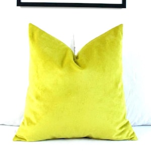 May include: A square throw cushion in a solid chartreuse yellow colour. The cushion features a soft, velvet-like texture with a visible seam along the edges, creating a structured shape. The fabric has a slight sheen that catches the light.