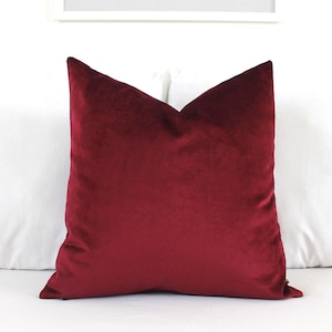 May include: A square throw pillow cover in a deep, rich burgundy red color. The fabric has a soft, short-pile velvet texture that catches the light, creating subtle variations in tone across the surface of the pillow cover.