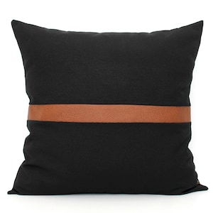 May include: Square black decorative pillow with a horizontal brown leather band across the center. The pillow has a soft, textured appearance and is set against a white background. This accent pillow is suitable for a sofa or bed.