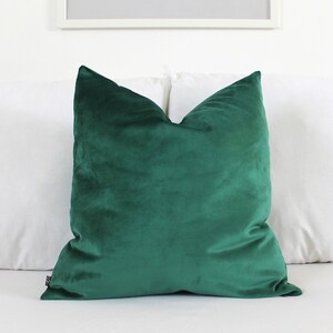 May include: A square throw cushion cover in a solid deep emerald green colour. The fabric has a soft, short-pile velvet texture with a subtle sheen. A small black rectangular brand tag is visible on the lower left side seam of the cushion cover.