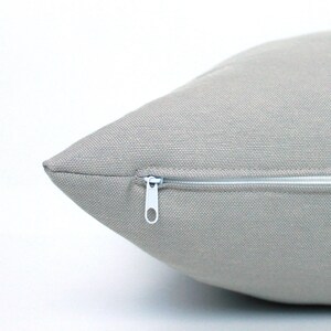 May include: Close-up of a light gray square pillow with a white zipper closure. The pillow has a textured fabric and a simple, minimalist design, suitable for home decor.