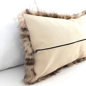 May include: A beige rectangular pillow with a faux fur trim in shades of brown and tan. The pillow has a black zipper closure on the back. The pillow is a decorative home accent.