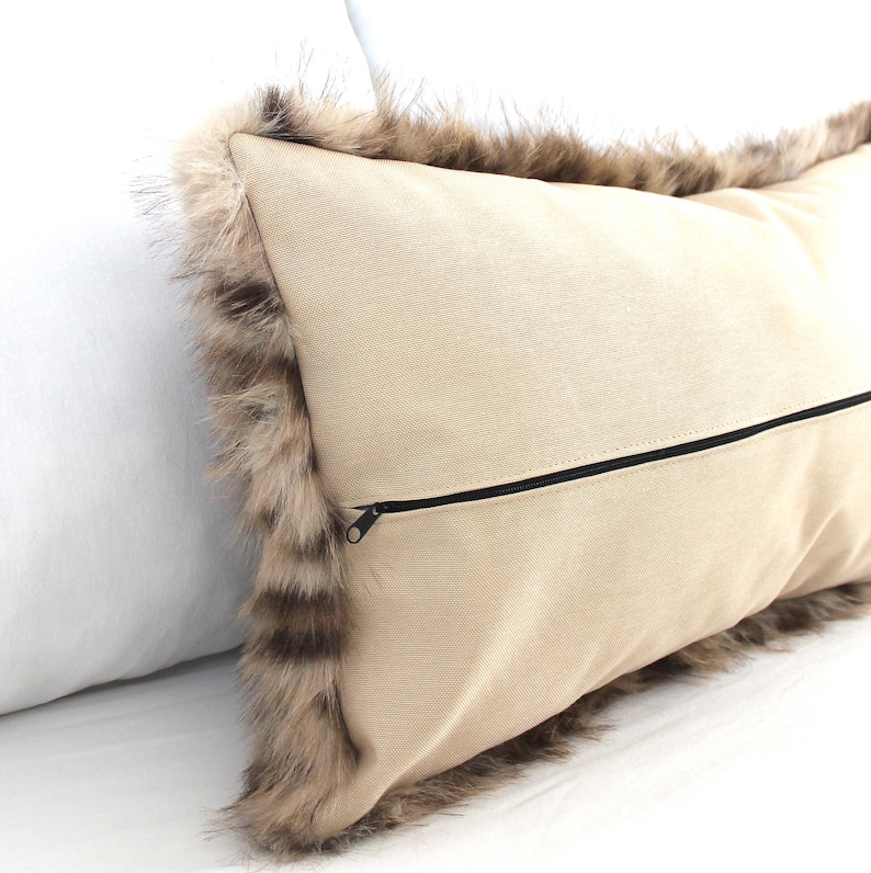 May include: A beige rectangular pillow with a faux fur trim in shades of brown and tan. The pillow has a black zipper closure on the back. The pillow is a decorative home accent.
