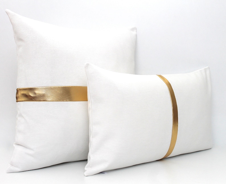 May include: Two white throw pillows with gold satin ribbon accents. The larger pillow has a horizontal ribbon, while the smaller pillow has a vertical ribbon.