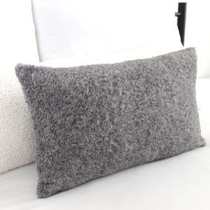 May include: A gray fluffy pillow with a soft, textured surface. The pillow is lying on a white bedspread.